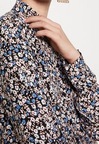 Blusa nera floreale con piccoli motivi di fiori blu, bianchi e rossi. Presenta un collo alto e polsini con bottoni. Tessuto dalla texture liscia.