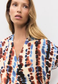 Ohlapna svilen top z abstraktnim vzorcem v rdeči, modri in črni barvi na kremni podlagi; ohlapen kroj z V-izrezom.