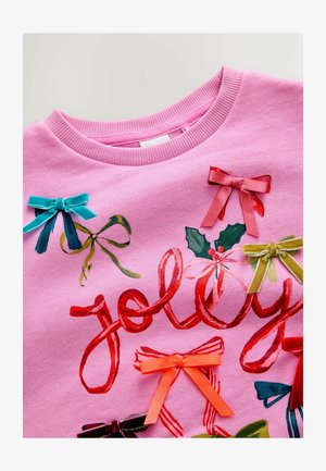 Roze sweatshirt met "Jolly" in het rood gedrukt, versierd met kleurrijke stoffen strikken in turquoise, groen, roze, oranje en paarse accenten.