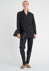 Musta bleiseri kaksirivisellä muotoilulla, yhdistettynä räätälöityihin mustiin housuihin ja teksturoituun mustaan laukkuun. Mallilla on päällään mustat sandaalit.