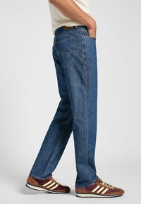 Jeans de mezclilla azul con un corte recto, cinco bolsillos y costuras amarillas. Combinados con zapatillas marrones y burdeos con detalles amarillos.