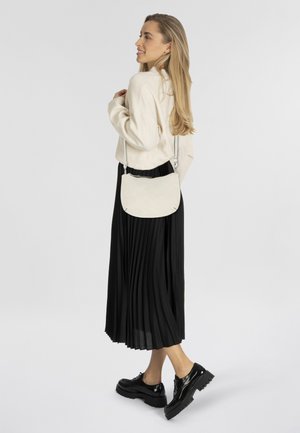 Sac en daim beige clair en bandoulière avec une forme incurvée, peu de coutures et une sangle réglable, associé à une jupe plissée noire et des chaussures brillantes.