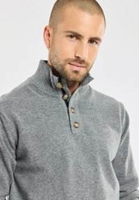 Pull en maille gris avec un col haut, comportant trois boutons. Le tissu texturé et le col côtelé accentuent le design.
