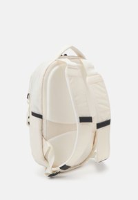 Beige reppu, jossa on pehmustettu muotoilu, säädettävät hihnat ja mustat yksityiskohdat. Ominaisuutena on jäntevä muoto ja sileä pinta.