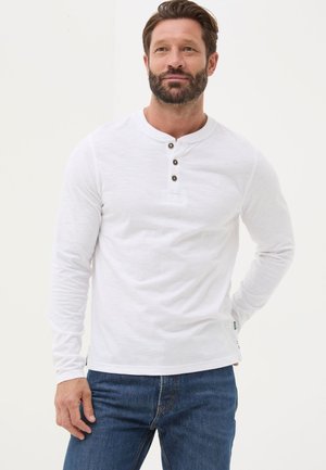 Man with beard wearing white long-sleeve henley shirt with three buttons and blue jeans, standing against a plain light background.