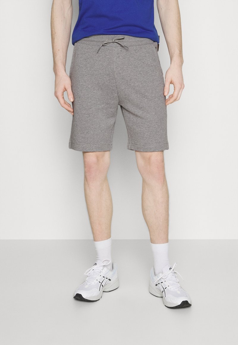 Homme portant un short gris à cordon, des chaussures de sport blanches, des chaussettes blanches et une chemise bleue, debout devant un fond clair uni.
