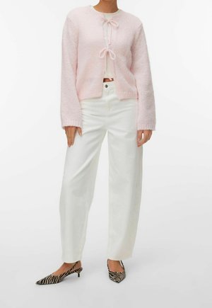 Femme portant un cardigan en maille rose clair avec des liens à l'avant, un pantalon blanc taille haute et des chaussures à talons bas imprimé zèbre.