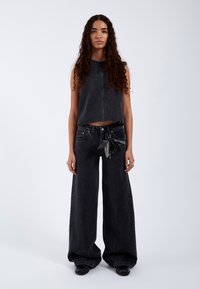 Vrouw met lang krullend haar, gekleed in een mouwloos zwart cropped topje en zwarte wijde jeans met een kettingaccessoire, staand tegen een witte achtergrond.