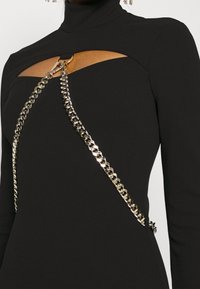 Robe noire à col montant avec un détail décolleté en forme de découpe et un fermoir en chaîne dorée traversant l'avant de l'épaule à la taille.