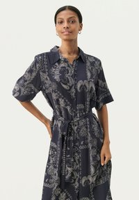 Rochie bleumarin cu un model paisley, mâneci scurte, închidere cu nasturi în față și un cordon asortat la talie. Material ușor, fluid.