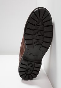 Zapato de cuero marrón con una suela de goma negra texturizada, que presenta profundos surcos y perforaciones decorativas a lo largo de los costados.