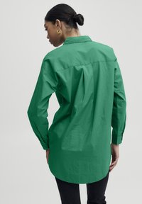 Green long-sleeve shirt with a collar, buttoned cuffs, and a back pleat design. Made of a smooth, lightweight fabric.