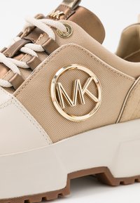 Baskets beiges avec une tige en tissu texturé, un logo accent doré et une semelle blanche épaisse avec une bande de roulement en caoutchouc brun. Les lacets sont blancs.