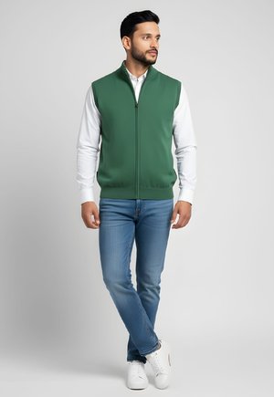Hombre de pie con las piernas cruzadas, vestido con chaleco verde con cremallera sobre camisa blanca, vaqueros azules y zapatillas blancas contra un fondo gris liso.