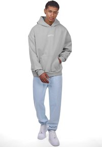 Grå hættejakke med kængurulomme og "DROPSIZE" logo. Sammen med lyseblå denimbukser og hvide sneakers. Glat stof, afslappet pasform.