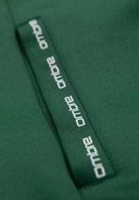 Un tessuto verde con una texture liscia presenta un logo "ombre" bianco prominente lungo una cucitura e dettagli di cucitura sottili.