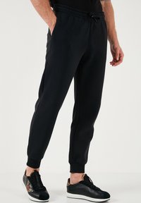 Schwarze Sweatpants mit elastischem Bund, Seitentaschen und schmal zulaufenden Knöcheln. Sie haben eine glatte Textur und werden mit schwarzen und goldakzentuierten Sneakers kombiniert.