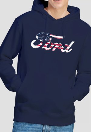 Mann trägt marineblauen Kapuzenpullover mit Ford-Logo, das mit Sternen und Streifen der amerikanischen Flagge gefüllt ist, Hände in den Vorderentaschen.
