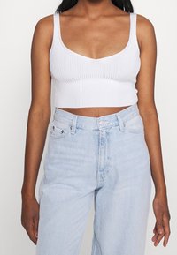 Crop top blanc côtelé avec un décolleté rond et des bretelles larges, associé à un jean en denim bleu clair taille haute avec une fermeture à boutons.