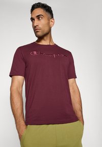 Bordeauxrotes Baumwoll-T-Shirt mit eingesticktem "Champion"-Logo in einem dunkleren Farbton. Kurze Ärmel und Rundhalsausschnitt. Modell trägt olivgrüne Hosen.