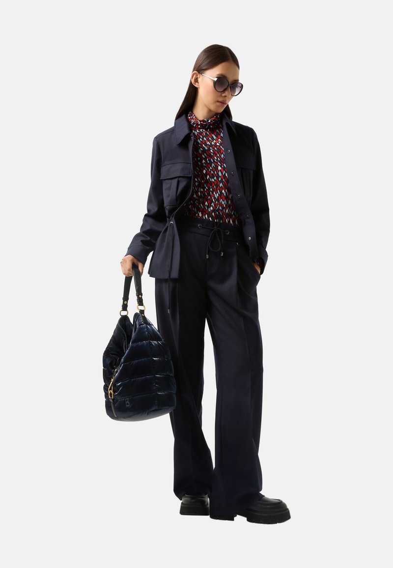 Marineblaues Jackett mit Taschen über einer gemusterten Bluse, kombiniert mit weitem Hosenbein und einer glänzenden, dunkelb blauen Puffertasche. Schwarze Plateauschuhe.