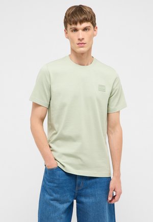 Junger Mann, der ein hellgrünes T-Shirt mit kurzen Ärmeln und blaue Jeans trägt, steht mit einer Hand in der Tasche vor einem einfarbigen grauen Hintergrund.