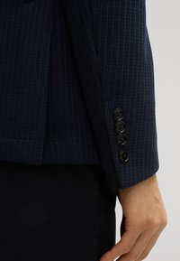 Donkerblauwe textuurjas met subtiele verticale strepen. Voorzien van vier zwarte knopen en een ventiele manchetdetail.