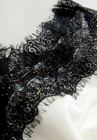 Melnas mežģīnes apdare ar sarežģītiem ziedu rakstiem un pērlītēm, kas kārtota virs gludas baltas auduma; delikāti frizēti mali.