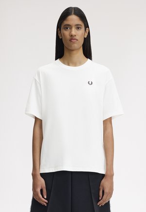 Ung kvinde med langt sort hår iført en enkel hvid t-shirt med korte ærmer og et lille sort laurbærkronelogo på venstre bryst.