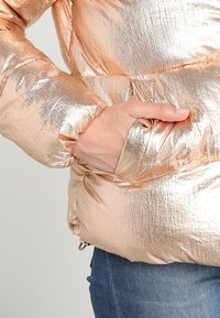 Ruka v kapse lesklé metalické růžovo-zlaté péřové bundy, nošené přes modré džíny, zaměřená na texturu bundy a detaily kapsy.