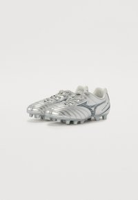 Le scarpe da calcio argento presentano una tomaia sintetica con un design testurizzato, lacci e tacchetti modellati per una migliore trazione. Il logo del marchio è visibile sul lato.
