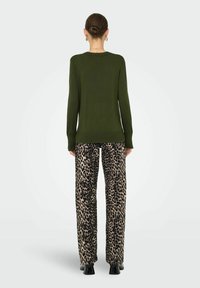 Grüner Strickpullover mit langen Ärmeln, kombiniert mit locker sitzenden, beigen Hosen mit einem schwarzen Leopardenmuster. Flache schwarze Schuhe.