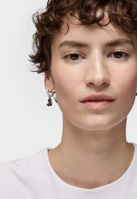 Boucle d'oreille en argent avec un charme noir en forme abstraite. Le modèle a les cheveux courts et bouclés et porte une chemise blanche simple. Peau nette et texturée.