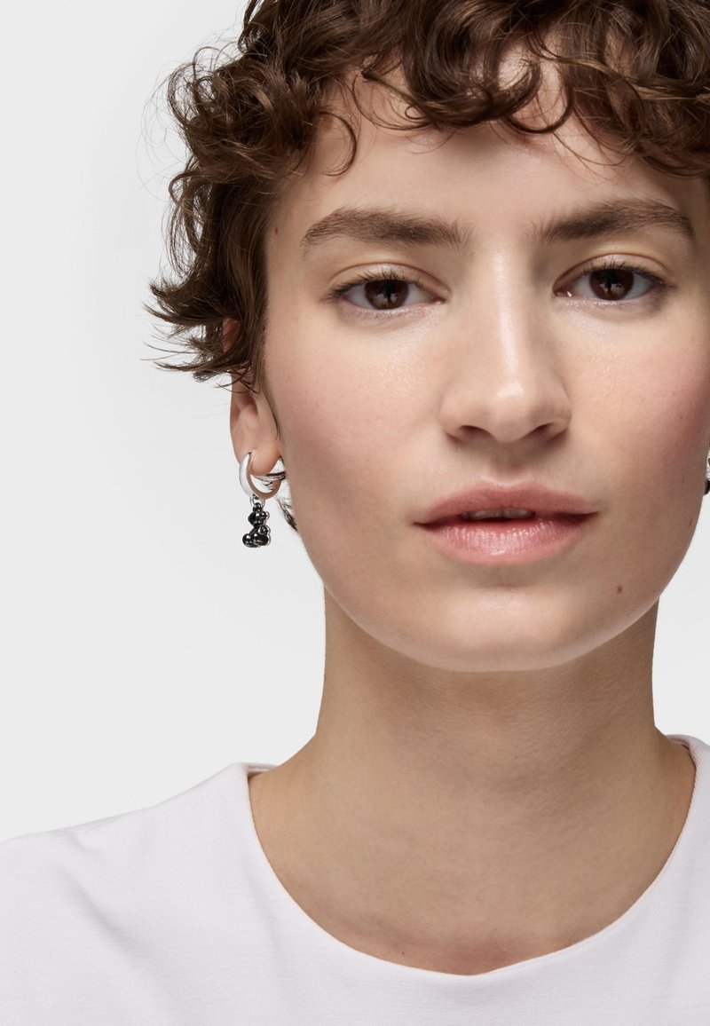 Boucle d'oreille en argent avec un charme noir en forme abstraite. Le modèle a les cheveux courts et bouclés et porte une chemise blanche simple. Peau nette et texturée.