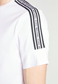 Wit T-shirt met korte mouwen met zwarte dubbele strepen en de tekst "MICHAEL KORS" herhaald langs de schoudernaad.