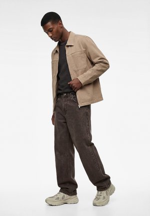 Mand iført beige jakke, sort tekstureret skjorte, mørke løstsiddende jeans og beige chunky sneakers, stående med hovedet let bøjet ned.