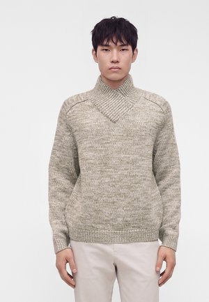 Jeune homme portant un pull en maille beige avec un col châle côtelé unique et un pantalon clair sur un fond uni.
