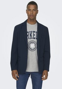 Marineblazer über einem grauen T-Shirt mit einem kreisförmigen Logo. Der Blazer hat einen schmalen Schnitt, Reverskragen und zwei Vordertaschen. Denim-Jeans vervollständigen das Outfit.