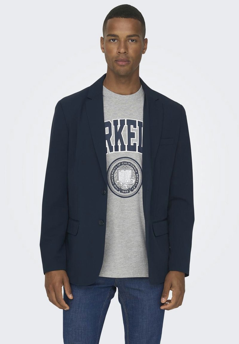 Marineblazer über einem grauen T-Shirt mit einem kreisförmigen Logo. Der Blazer hat einen schmalen Schnitt, Reverskragen und zwei Vordertaschen. Denim-Jeans vervollständigen das Outfit.