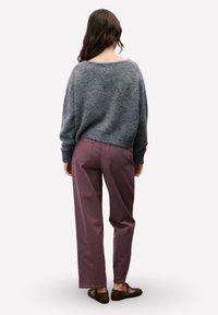 Vrouw met lang golvend haar, gekleed in een losse grijze trui, mauve rechte broek en donkerbruine platte schoenen, staand met haar gezicht naar buiten gekeerd.