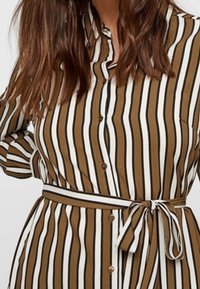 Camicia a righe marroni e bianche con bottoni, vita con cintura e colletto classico. Il tessuto appare liscio con un design leggermente strutturato.