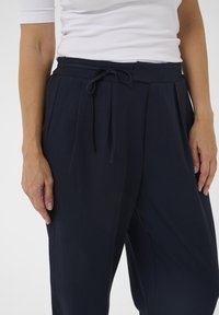 Zelo bližnji posnetek osebe, ki nosi modro visoko pasovne hlače z zavezanim pasom in belo rebrasto majico s kratkimi rokavi.