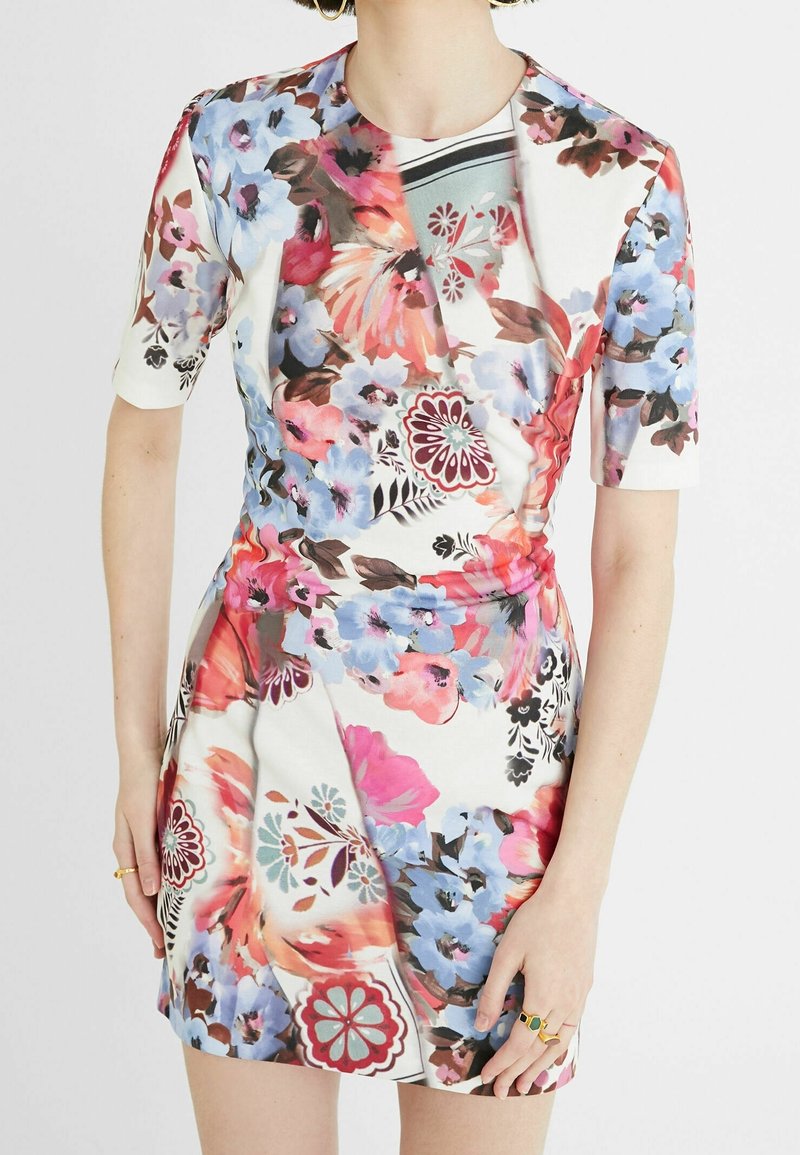 Femme portant une robe blanche à manches courtes avec des motifs floraux roses, bleus et marron et un détail froncé à la taille.