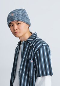 Πλεκτό μπλε σκουφί με ραβδωτή υφή, φορεμένο με ένα πουκάμισο κοντομάνικο ριγέ που διαθέτει ναυτικό, ανοιχτό μπλε και λευκές ρίγες.