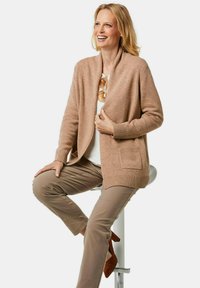 Beige gebreide cardigan met een sjaalkraag en geribbelde manchetten, voorzien van een voorkantzak. Draagt over een witte top en gecombineerd met lichtbruine broek.