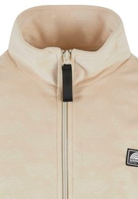 Vaaleanbeige fleece-takki, jossa on korkea kaulus, etuvetoketju ja musta vetoketjun vetimen. Sisältää hienovaraisen kuvioidun tekstuurin ja logopatchin sivussa.