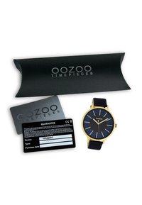 Montre avec bracelet en silicone noir, boîtier doré, cadran bleu étoilé et index horaires dorés. Comprend un emballage noir et des cartes de garantie.