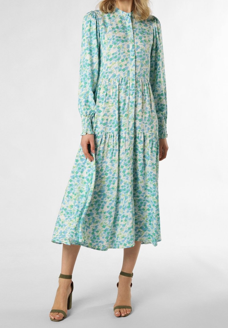 Femme portant une robe à manches longues, longue jusqu'aux chevilles, avec un motif abstrait bleu, vert et blanc, et des sandales à talons hauts vert olive.
