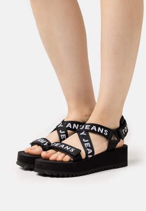 Sandales à plateforme noires avec des sangles en tissu portant le texte "TOMMY JEANS". Semelle en caoutchouc épaisse avec texture striée, design à bout ouvert.