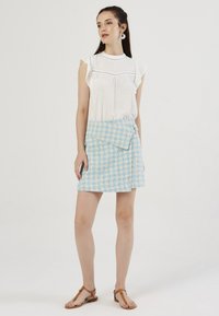 Witte blouse met stippen en ruches aan de mouwen, gecombineerd met een blauw-geel geruite wikkelrok met een zijzak en een gespdetail.